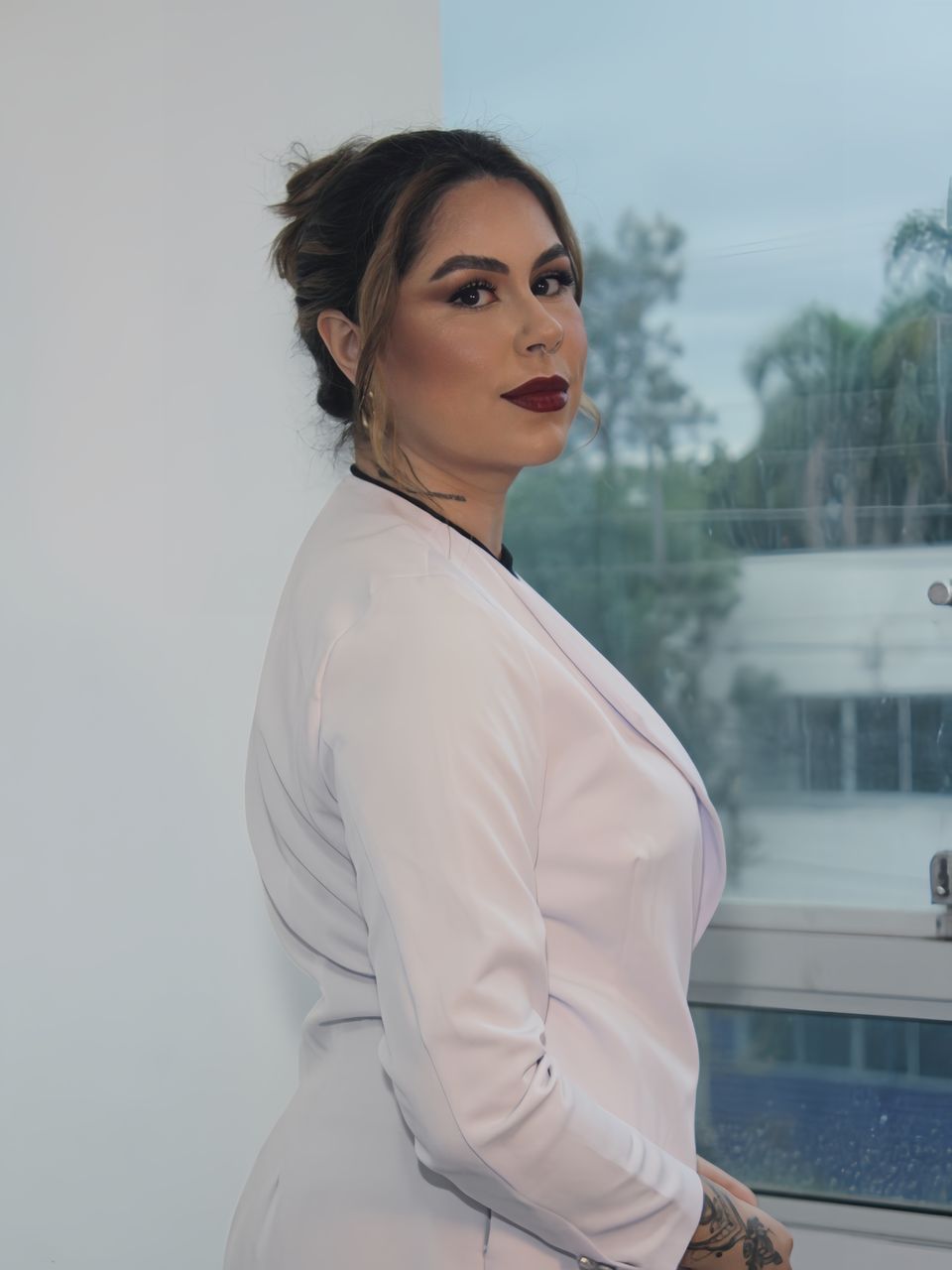 Evelyn Soares, de jaleco branco, em pé diante da janela do consultório.